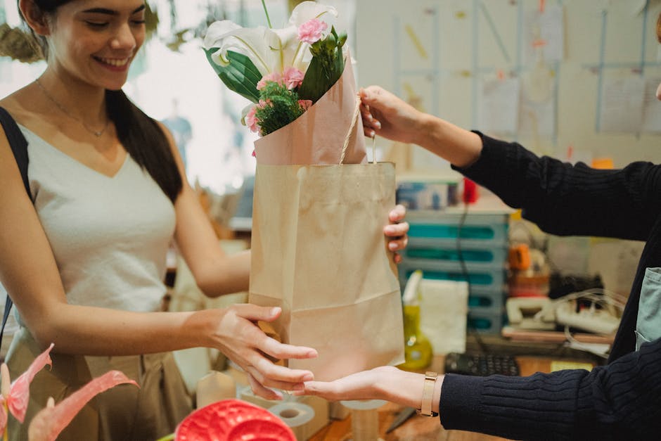 A woman with long dark hair, wearing a white sleeveless top, is smiling as she receives a bouquet of fresh flowers wrapped in pink and beige paper, presented in a light brown paper bag from another pe