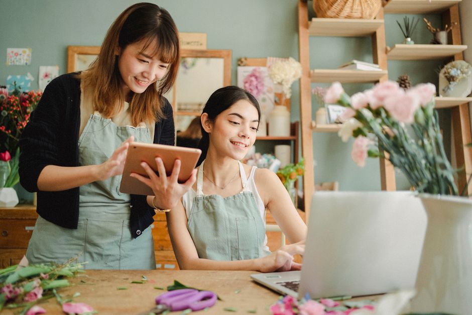 Two women in a flower shop working on a floral arrangement. The woman sitting at the wooden table has dark hair and is wearing a light grey apron, smiling as she looks at her laptop. The standing woma