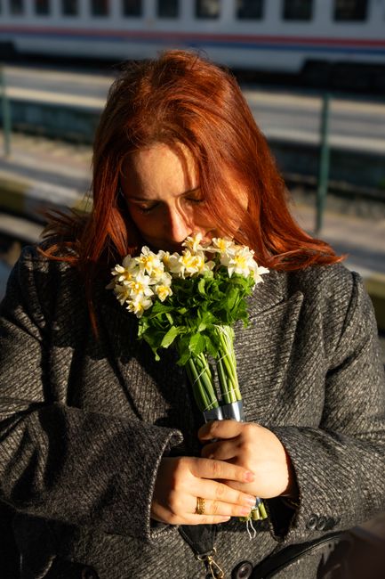 A woman with red hair, wearing a dark grey textured coat, is holding a bouquet of fresh yellow and white daffodils with green stems and leaves, wrapped in clear plastic, close to her face as she appea
