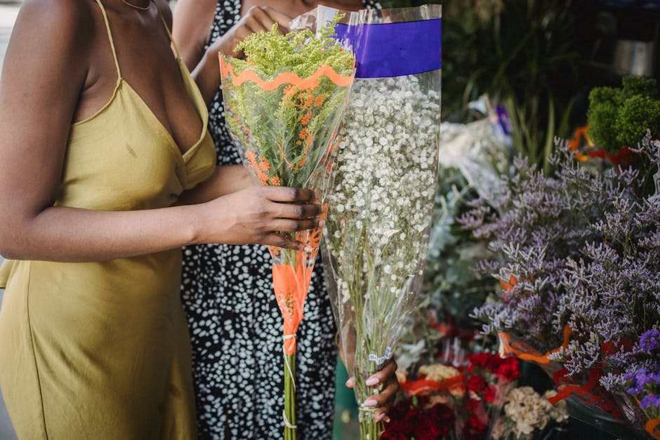 A woman dressed in a yellow satin slip dress is holding a bouquet of green and purple flowers wrapped in clear and orange plastic wrapping, while standing in a flower shop. Next to her, another person