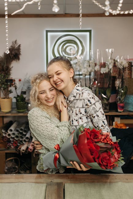 Two young women with light skin and curly blonde hair sharing a joyful moment inside a flower shop. One woman, dressed in a light green, floral-patterned top, is smiling brightly, while the other, wea