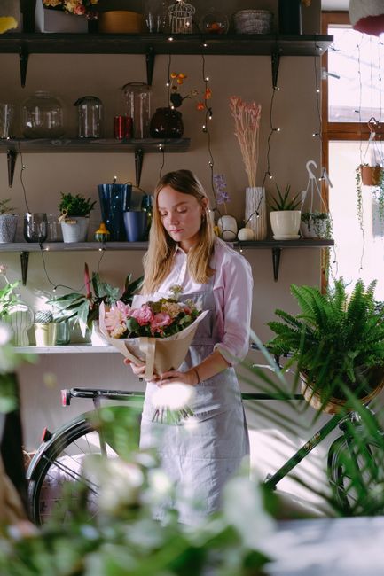 A young woman with light brown hair, dressed in a white shirt and apron, is standing indoors in a well-lit florist shop, holding a bouquet of fresh pink roses, white peonies, and greenery wrapped in b