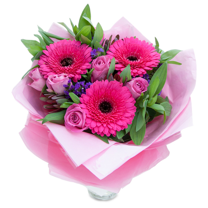 The charm of this arrangement lies in its contrast of bold and gentle notes: three fuchsia Gerbera daisies burst forward with saturated colour and sculptural petal layers, their dark centres grounding the palette. Surrounding them, roses in a soft lavender-pink or dusty rose hue unfurl with delicate, velvety petals, creating pockets of tender texture. Between these larger blossoms, clusters of purple statice add a light, airy punctuation - a fine, papery texture that seems to float among the stems and gives the bouquet a casual, almost wildflower feel. A generous framing of green leaves, likely salal or eucalyptus, provides weight and a fresh scent, their cool green tones amplifying the pinks and lending an organic fullness. The wrapping is deliberately elegant: a matte pale pink tissue inner wrap cushions the blooms while a sheer fuchsia-pink cellophane overlay, slightly crinkled, imparts a soft shimmer and keeps the colour story unified. Careful folding around the base suggests the work of an attentive hand, and the visible rim of a clear glass vase at the bottom hints that the flowers are arranged for immediate display. I imagine placing this on a windowsill with light from Well Hall Pleasaunce slanting in, the bouquet filling a room with gentle rose perfume and a bright, optimistic energy - perfect for celebrating a new home, cheering a friend, or marking a special birthday in Eltham.