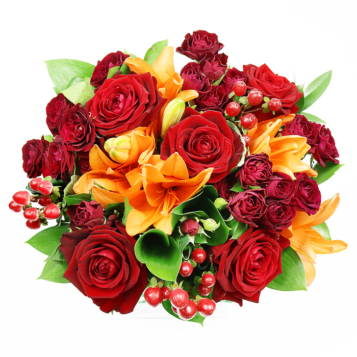 From a direct overhead angle on a pristine white backdrop, this sumptuous bouquet unfolds like a velvet mosaic of reds, oranges, and greens, meticulously arranged by a local Eltham hand. Several large, fully opened deep red roses command the centre with their layered, velvety petals, each bloom reading as a quiet declaration of love and tradition. Bright orange Asiatic lilies are woven through the composition, their trumpet silhouettes and visible stamens creating upward energy while a few buds sit mid-unfurl, promising continued freshness. Tucked in among these are tight clusters of dark red spray roses, compact and plush, which add concentrated depth and a jewel-toned shadow beneath the main flowers. Glossy hypericum berries pepper the bouquet in small groups, their round, reflective surfaces adding lively accents and a hint of festive cheer. Deep green leaves - the broad, glossy types we commonly use in Eltham arrangements, such as ruscus or salal - peek and arc around the flowers, lifting the warm palette and providing a crisp botanical frame. The lighting is gentle and even, chosen to reveal both petal texture and berry gloss without distraction. This composition evokes autumnal warmth and romantic intent, ideal for anniversaries, celebratory gestures, or simply brightening a home near Eltham Palace or Avery Hill Park. Crafted with the same care Eltham Florist brings to every hand-tied piece, the bouquet reads as both abundant and thoughtfully balanced, its circular form and dense packing promising a full-bodied scent and an immediate visual presence on a sideboard or dining table.