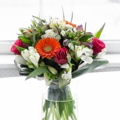 This masterfully arranged bouquet offers a harmonious blend of colour and form, perfect for brightening homes from Well Hall Road to Avery Hill. Creamy peach gerberas with deep brown centers create a soft halo, inviting the eye inward to fiery orange counterparts that pulse with energy, reminiscent of Eltham's vibrant market days. Among these, tight magenta roses add depth and romance, their velvety petals shimmering against a scattering of white alstroemeria and petite spray roses, which infuse the bouquet with lightness and refined texture. Interwoven among the focal flowers are slender, pointed greens and airy white fillers, contributing to a sense of abundance and easy movement. The distinctive base-broad, lush aspidistra leaves-as if hugging the blooms, grounds the stunning display, echoing the tranquility of Eltham Palace gardens. Presented in a tall, clear vase, the arrangement captures the interplay of organic stems and vivid flowers, accentuated by the filtered daylight streaming through a nearby window. The scent, gentle and rejuvenating, hints at both rose sweetness and fresh greenery. Ideal for joyous celebrations, brunches with friends, or heartfelt tokens marking new beginnings, this bouquet from a trusted Eltham florist is more than a decorative piece; it sings with thoughtful intention and local pride, expertly balancing every petal, leaf, and hue for an uplifting experience.